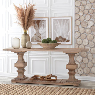 A rustic wooden console table with turned legs and a slatted base, styled with vases, pampas grass, a bowl of greenery, and a decorative chain on a woven rug in a bright interior setting.