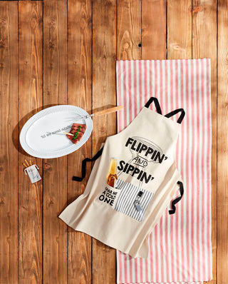 Canvas grilling apron gift set styled on a wooden table with a striped runner, serving platter, and grilling accessories