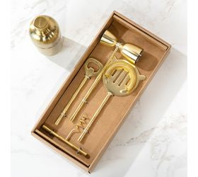 A set of gold-colored metal bar tools, including a jigger, spoon, and strainer, presented in a brown cardboard tray with a matching gold tumbler on a white marble surface.