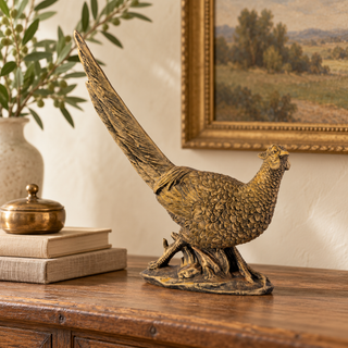 A decorative bronze-finished pheasant figurine displayed on a wooden console table in a styled room setting with books, a vase, and framed wall art.
