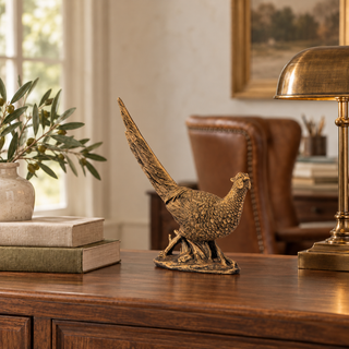 An ornate bronze-finished pheasant figurine displayed on a dark wood console table in a warm, elegant room setting with a leather armchair, brass lamp, and books in the background.