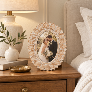 Whitewashed wood oval 4x6 photo frame with carved autumn leaves displaying a newlywed couple on a warm wood nightstand beside neutral bedding, a ceramic lamp, and a brass ring dish.
