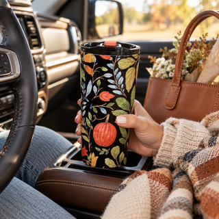 20 oz insulated stainless steel coffee tumbler featuring watercolor pumpkins and fall florals on a black background, held inside a cozy car with a cream knit sweater, plaid blanket, and brown leather tote.
