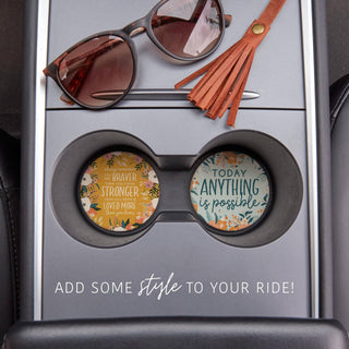 Two inspirational floral ceramic coasters displayed inside vehicle cup holders beside sunglasses and a leather tassel.