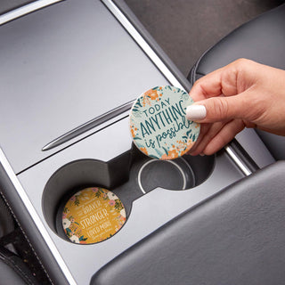 Hand removing the “Today Anything Is Possible” ceramic car coaster while the coordinating floral coaster remains in the adjacent cup holder.