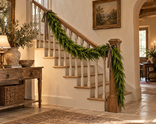 72-inch Real Touch cedar garland draped along an interior wood staircase railing with muted velvet ribbon in a warm European cottage entryway.