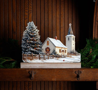 Snowy town church Christmas silhouette displayed unlit on a wood mantel with an evergreen tree, fence, wreath, and steeple.