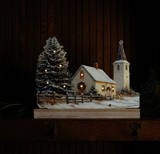 Lighted snowy town church Christmas silhouette glowing warmly on a dark wood mantel with evergreen accents.