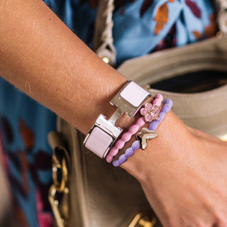 A close-up of a person's wrist wearing a stack of bracelets, including a wide silver-toned bracelet with a pale pink rectangular inlay, two thin elastic bracelets in pink and lavender, and a small silver butterfly charm.