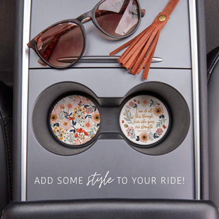 Two colorful floral Christian ceramic coasters displayed inside vehicle cup holders beside sunglasses and a leather tassel.