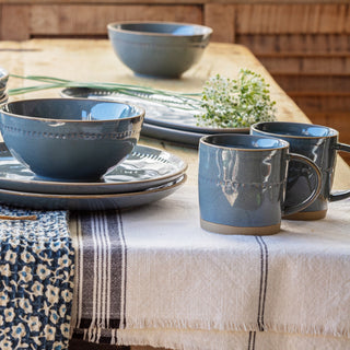 A set of rustic blue-gray ceramic tableware, including bowls, plates, and mugs with a glossy finish and gold-toned trim, arranged on a table covered with a cream tablecloth and a patterned runner.