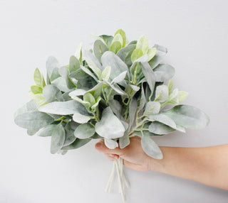 A hand holding a bouquet of artificial eucalyptus-style greenery with silvery gray leaves and lighter sage-green foliage against a plain light background.