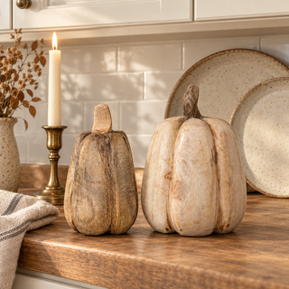 Small and large handmade mango wood pumpkins styled on a warm kitchen counter with neutral stoneware, an aged-brass candlestick, and oatmeal linen.