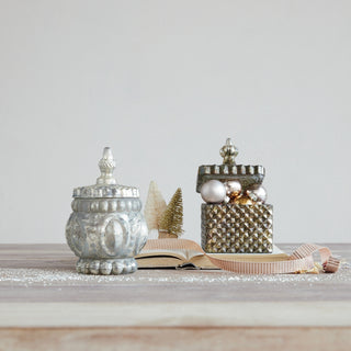 Two decorative galvanized metal lidded boxes with ribbed and textured surfaces, styled on a light wood surface with a ribbon, miniature trees, and ornaments against a neutral background.