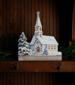 Snow-covered Christmas church displayed on a rustic wooden mantel with evergreen branches and dark wood paneling.
