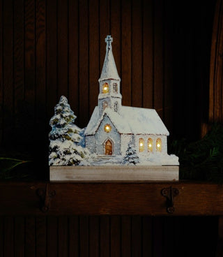 Illuminated snowy Christmas church with warmly glowing windows styled on a rustic mantel against dark wood.