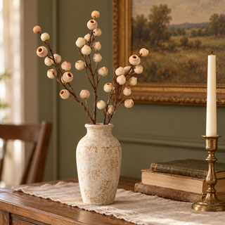 Peach eucalyptus pod sprays arranged in a weathered cream vase on a warm wood console with vintage books, aged brass, and a muted green wall.