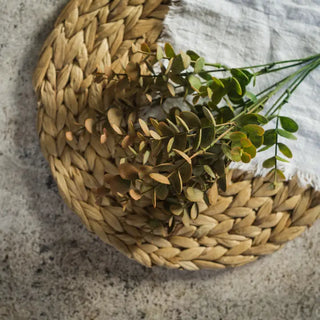 A circular woven straw placemat in a natural tan color, styled with a dried green leafy sprig and a corner of white textured fabric on a gray stone surface.