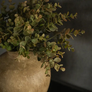 Artificial green eucalyptus branches with round leaves arranged in a rustic beige ceramic vase against a dark gray background.