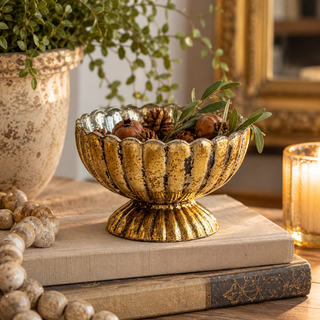 Antique gold mercury glass pedestal bowl with scalloped edges, filled with decorative pomegranates, pinecones, and greenery, styled on vintage books beside a rustic planter and glowing candle.