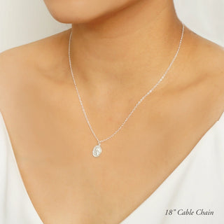 A close-up of a model wearing a delicate silver cable chain necklace with a small oval clear pendant. The model is wearing a white top, and the background is a soft, neutral light gray.