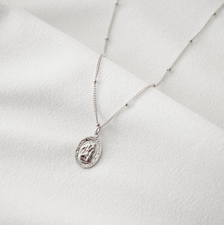 A silver chain necklace with an oval Saint Miriam medal pendant, displayed on a textured white fabric background.