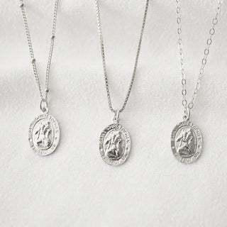 Three silver Saint Christopher medal necklaces with round medallion pendants on delicate chains, displayed against a soft white textured background.