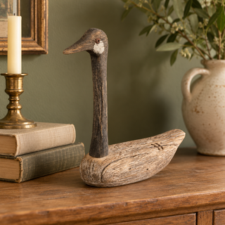 Rustic wooden goose sitter styled on a warm wood console with vintage books, an aged-brass candlestick, and a cream ceramic vessel against a muted olive wall.